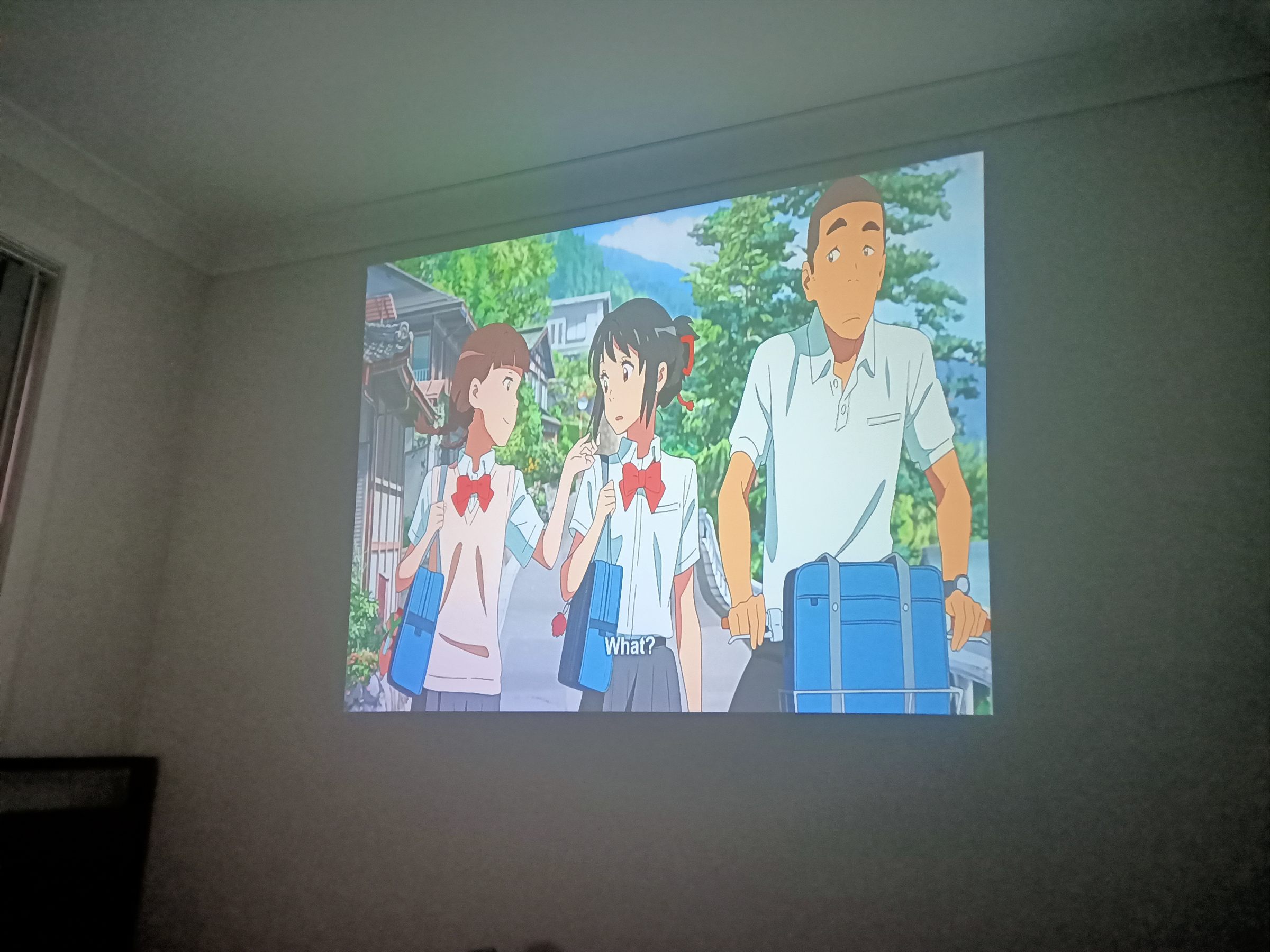 A scene from Your Name projected onto a bedroom wall — two schoolgirls and a man with a suitcase on a village street