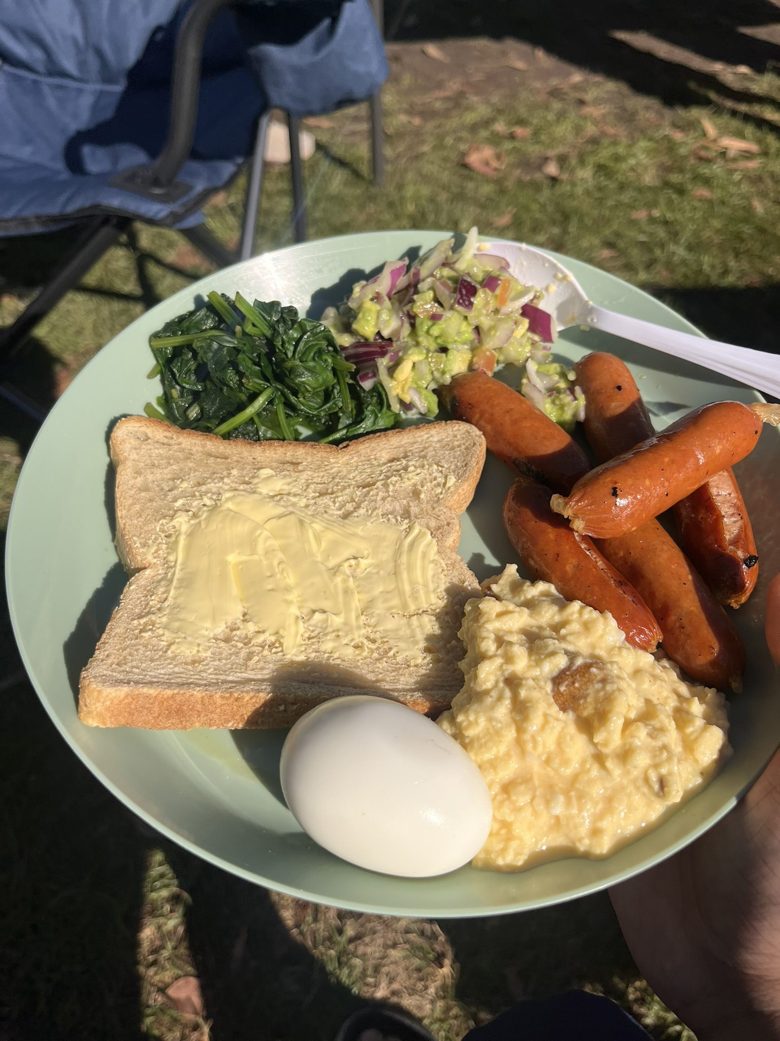 A camp breakfast on a pale green plate — buttered toast, wilted spinach, avocado and red onion salsa, grilled chipolatas, scrambled egg and a peeled boiled egg — held over grass beside a camping chair