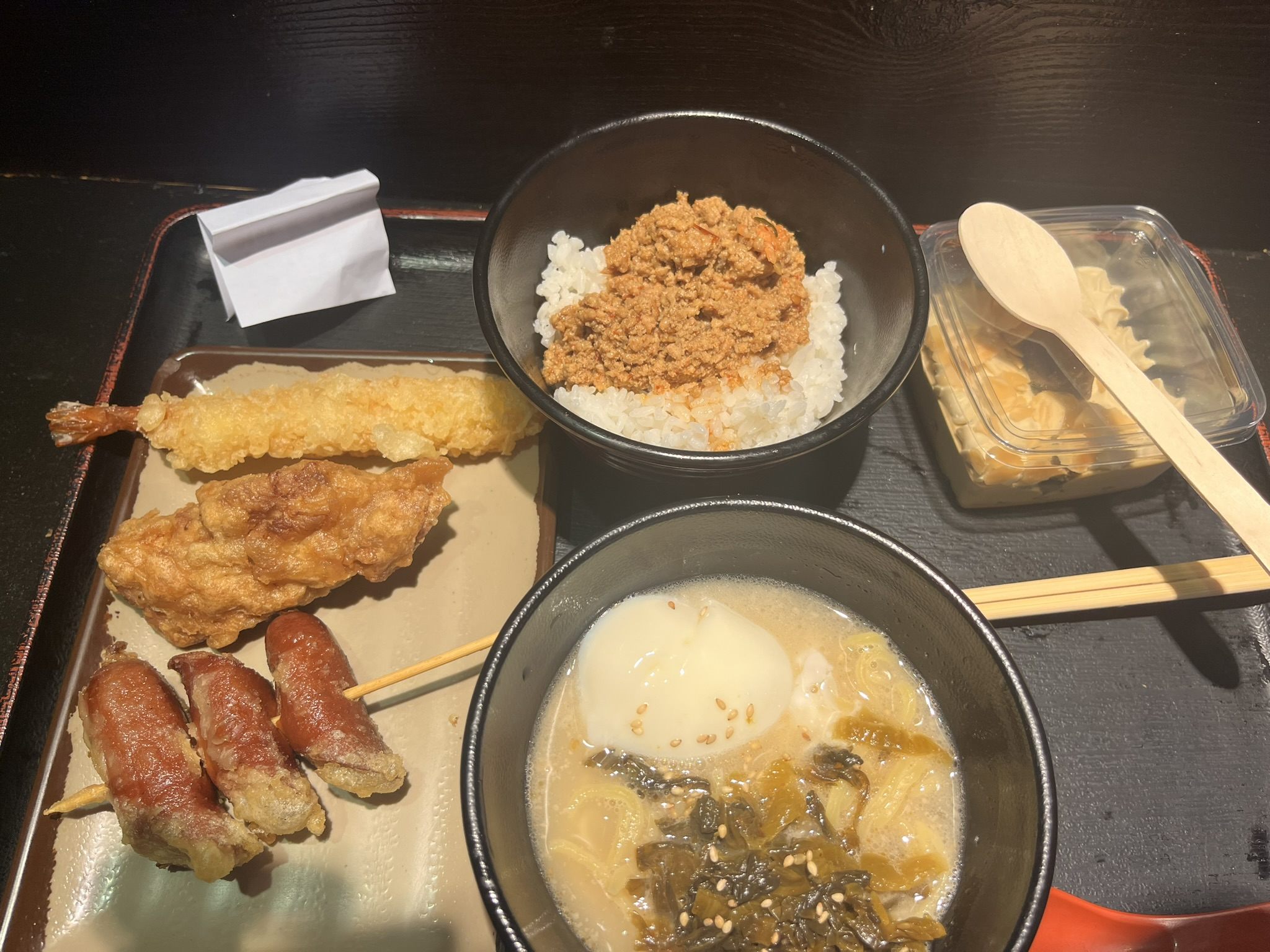 A Japanese set meal on a black tray: prawn tempura, fried chicken and sausage skewers, a rice bowl topped with minced meat, and a bowl of soup with a soft egg and pickled greens