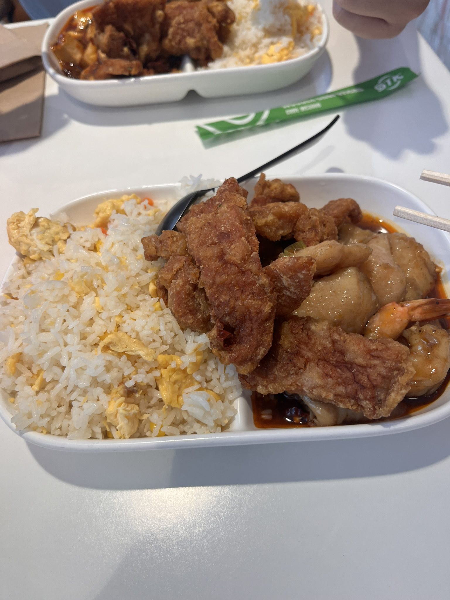 A takeaway tray of egg fried rice with fried chicken, glazed chicken and a prawn in dark sauce, on a white table