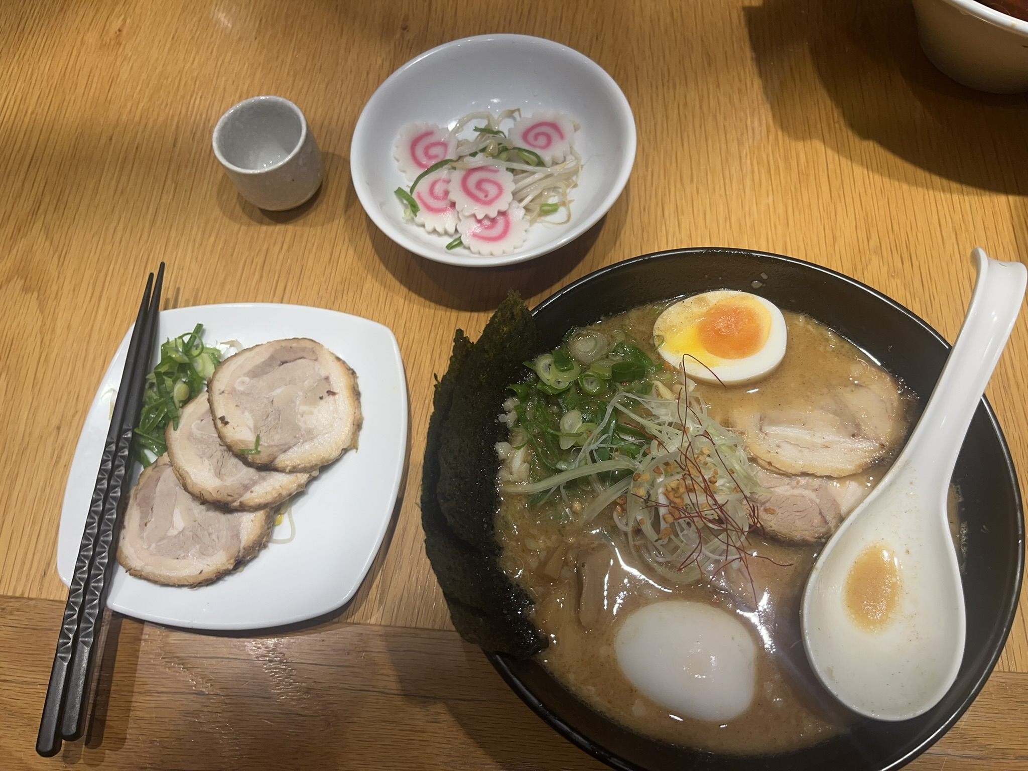 A ramen set on light wood: a black bowl with chashu, nori and spring onion, a side plate of sliced chashu, and a small bowl of narutomaki and bean sprouts