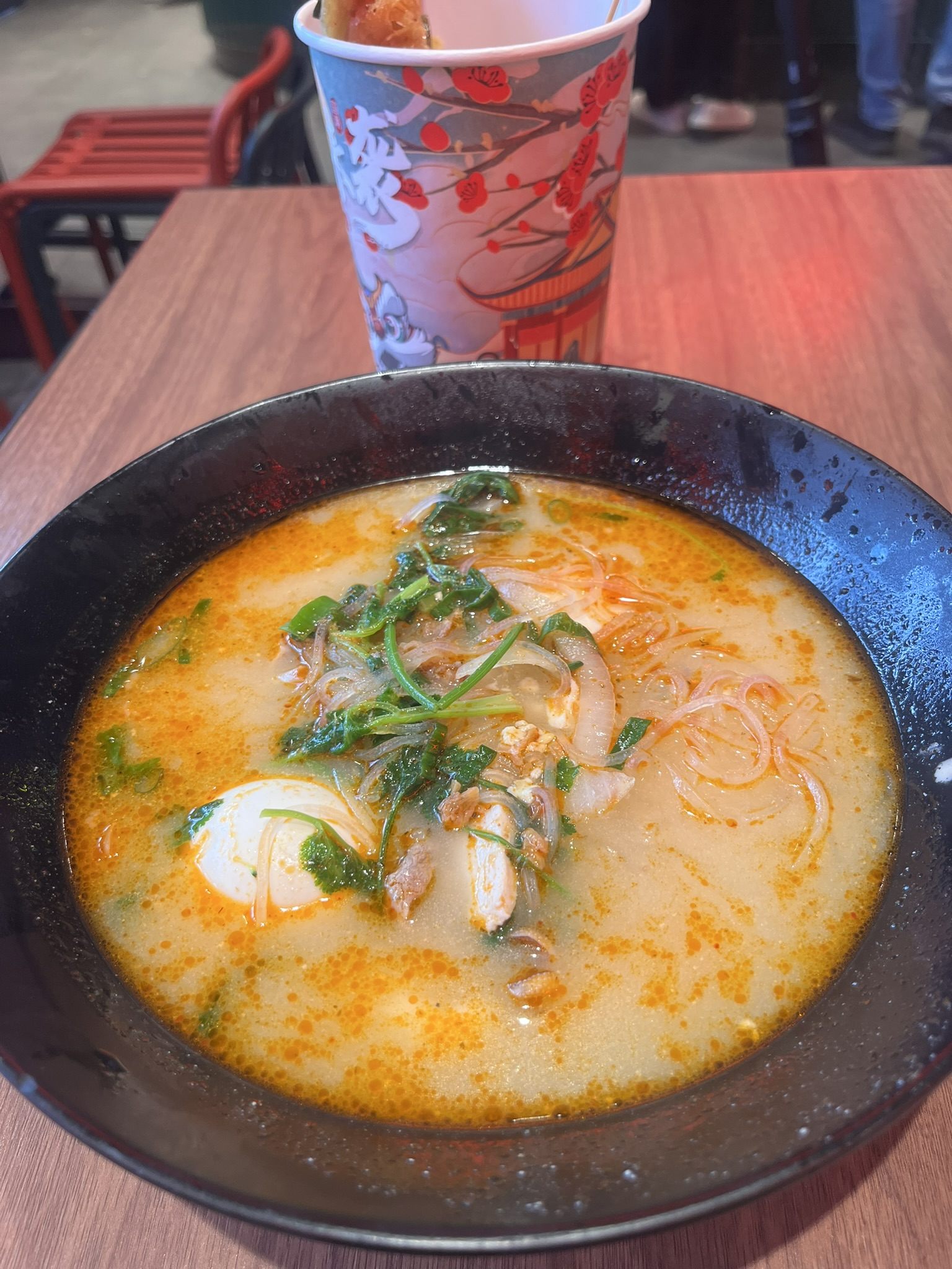 A black bowl of soup noodles with greens, sliced chicken and egg, on a wooden food-court table
