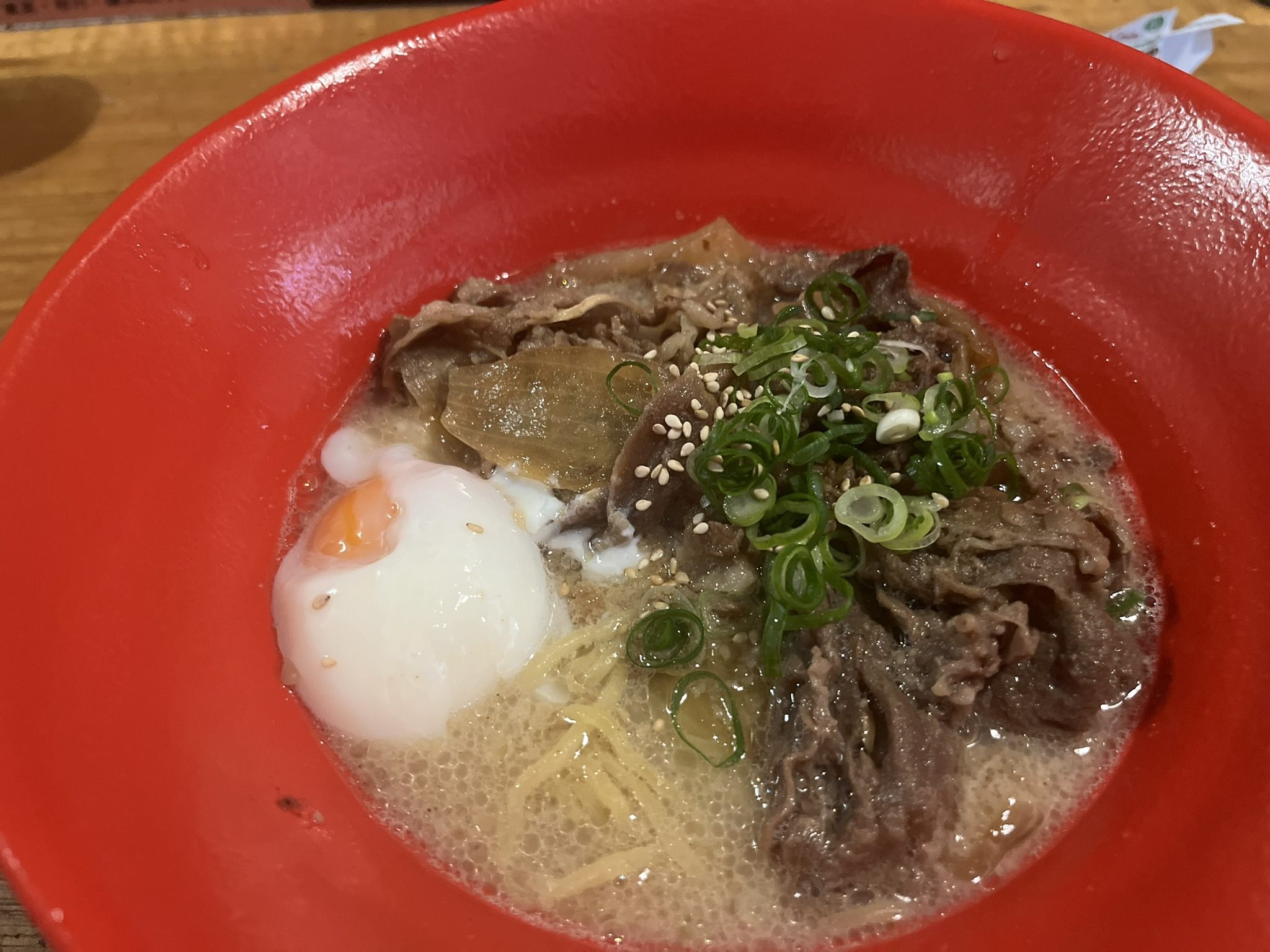 A red bowl of udon with sliced beef, spring onion, sesame seeds and a soft poached egg