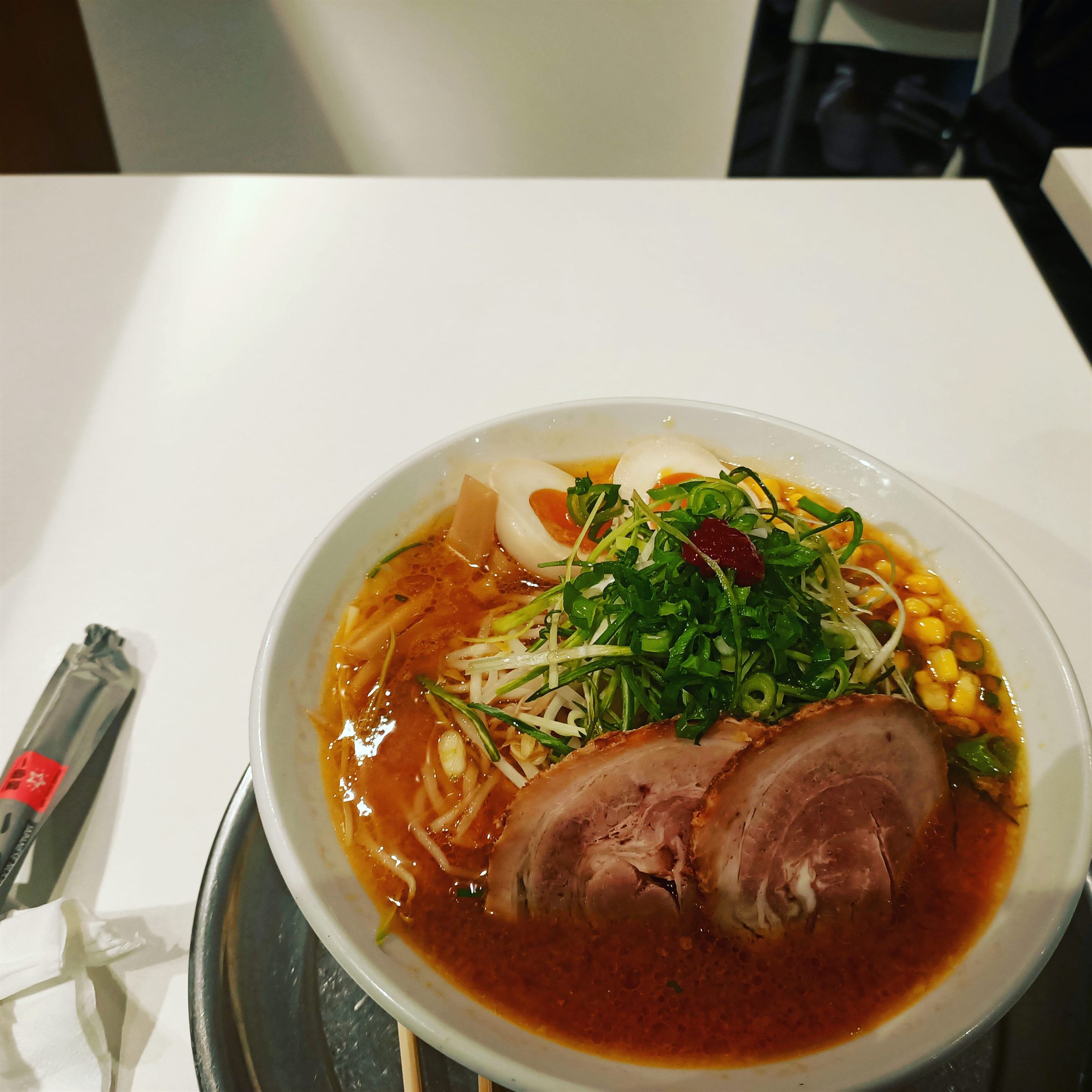 A white bowl of spicy miso ramen with two thick slices of chashu, sweetcorn, bean sprouts and greens
