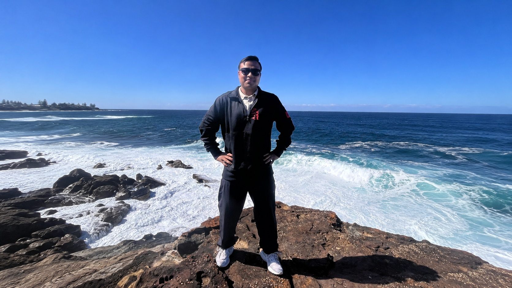 Standing on dark coastal rocks with surf breaking behind, under a clear blue sky