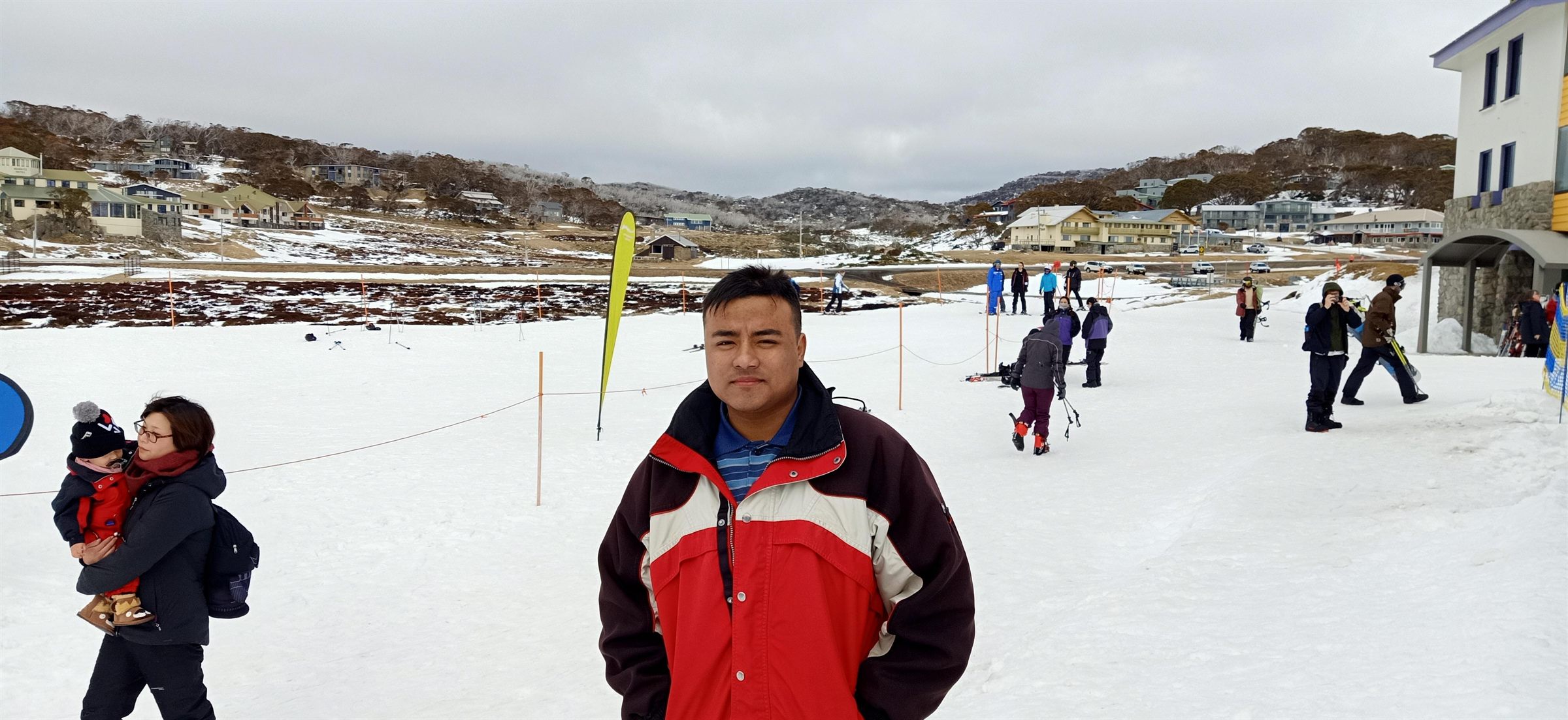 Standing in a red and cream jacket on packed snow, with a snow-covered village, bare gum trees and low hills behind