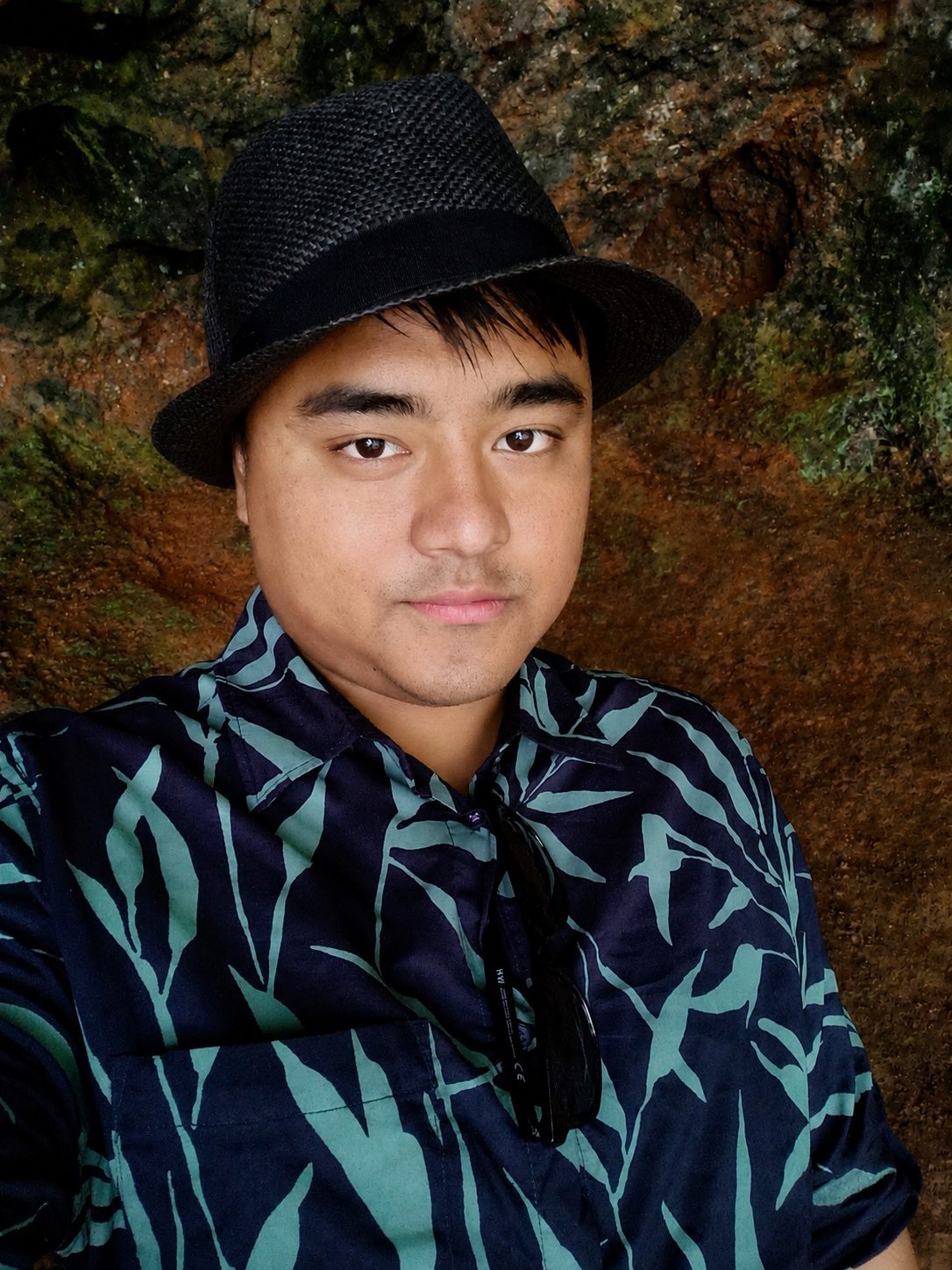 Portrait of Rijan Pradhan in a black hat, against a mossy rock face