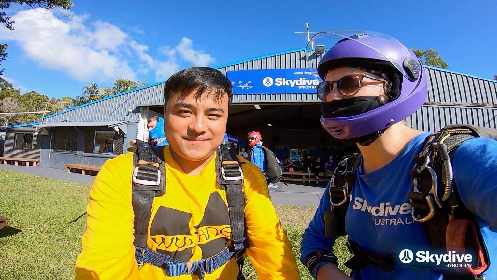 Standing on grass in a skydiving harness outside the Skydive Byron Bay hangar under a clear sky, beside a tandem instructor in a purple helmet